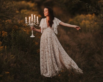 May include: A woman in a white lace dress holds a candelabra with lit candles in a field of tall grass.