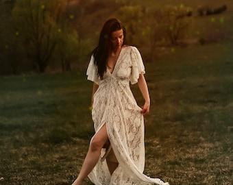 May include: A woman in a white lace dress walks through a field of green grass. The dress has a deep V-neckline and a flowing skirt. The woman's hair is long and dark.
