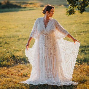May include: An ivory lace wedding dress with a V-neck and flowing sleeves. The dress has intricate floral patterns and a scalloped hem. The bride is standing in a sunlit field.