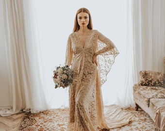 May include: A woman in a long, flowing, beige lace dress with a bouquet of white and green flowers. She is standing on a patterned rug in a room with white curtains and a chandelier.