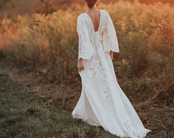 Puede incluir: Vestido de novia de encaje blanco con mangas largas y cola fluida. El vestido presenta una espalda en V profunda y se usa en un campo de hierba alta al atardecer. Estilo bohemio.