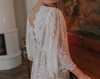 May include: A white lace wedding dress with long sleeves and a low back. The dress has a fitted bodice and a flowing skirt.