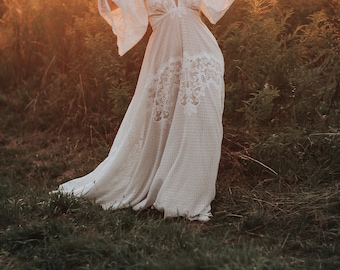 May include: A woman in a white lace dress stands in a field of tall grass. The sun is setting behind her, casting a warm glow on the scene.