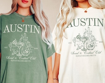 May include: Two oversized t-shirts with a cactus and cocktail graphic. The text on the shirts reads "AUSTIN EST. 2024 Social & Cocktail Club OLIVIA'S BACHELORETTE". The shirts are in moss green and ivory.