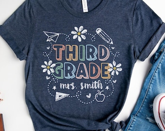 Può includere: T-shirt blu navy con la scritta "THIRD GRADE mrs. smith" in un design colorato e stravagante. La maglietta presenta un aeroplano di carta, una matita, fiori e una grafica a forma di mela. La maglietta è abbinata a jeans blu e scarpe da ginnastica bianche.