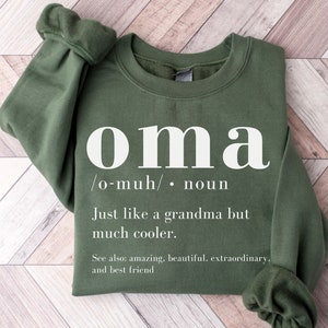 May include: Military green sweatshirt with the word "oma" in large white letters. The text below defines "oma" as "Just like a grandma but much cooler." Additional text includes "See also: amazing, beautiful, extraordinary, and best friend."