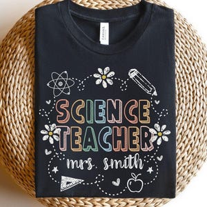 May include: Black t-shirt with the words "SCIENCE TEACHER mrs. smith" in colorful, outlined letters. The design includes science-themed doodles like an atom, pencil, flowers, and an apple. The shirt is displayed on a woven surface.