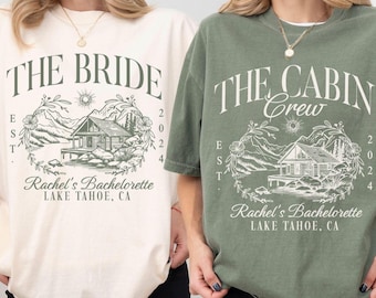 Puede incluir: Dos camisetas, una marfil y otra verde musgo, con los textos "THE BRIDE" y "THE CABIN CREW" impresos. Cada camiseta presenta un gráfico de montaña y cabaña con el texto "Rachel's Bachelorette LAKE TAHOE, CA".