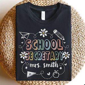 Peut inclure: T-shirt noir avec l'inscription "SCHOOL SECRETARY mrs. smith" en lettres color&eacute;es et soulign&eacute;es. Le motif comprend un avion en papier, un crayon, des fleurs et une pomme. Le t-shirt est pr&eacute;sent&eacute; sur une surface tiss&eacute;e.