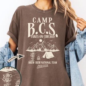 May include: Espresso-colored t-shirt with the text "CAMP B.C.S. TAKES ON CHICAGO." A graphic of mountains, a tent, and a campfire is below the text. The shirt also says "BREW CREW NATIONAL TEAM JULY 2026." A small graphic shows a signpost with "CAMP B.C.S. 2026 THIS WAY."