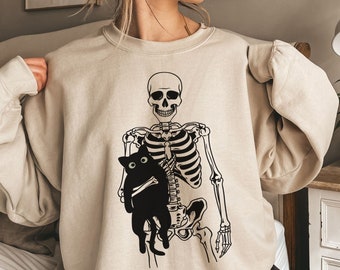 May include: A beige sweatshirt with a black skeleton holding a black cat. The skeleton is wearing a black shirt and the cat has white eyes.