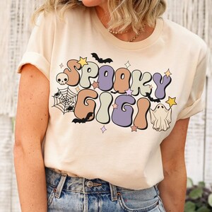 May include: A cream coloured t-shirt with a retro-style graphic that says "Spooky Gigi" in colourful letters. The graphic also includes a ghost, bats, and a spider web.