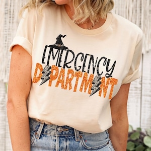 May include: A white t-shirt with a black witch hat and orange glitter text that reads "Emergency Department".