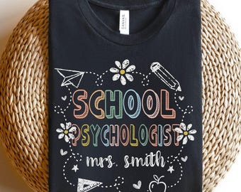 May include: Black t-shirt with the words "SCHOOL PSYCHOLOGIST mrs. smith" in colorful, outlined letters. The design includes a paper airplane, pencil, flowers, stars, an apple, and a ruler.