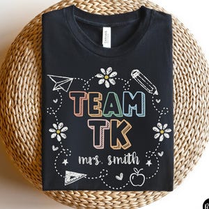 May include: Black t-shirt with the text "TEAM TK" in colorful letters, with the name "mrs. smith" below. The design includes a paper airplane, pencil, flowers, and an apple. The shirt is displayed on a woven surface.