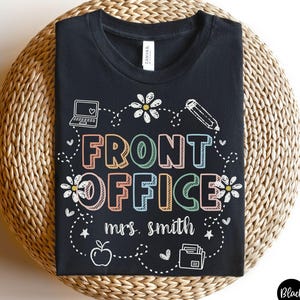 May include: Black t-shirt with the words "FRONT OFFICE" in colorful, block letters. The design includes a laptop, pencil, flowers, and an apple. The name "mrs. smith" is printed below the text.