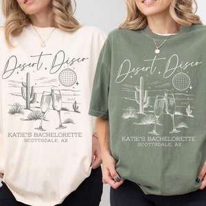 May include: Two t-shirts with a desert disco design. The shirts are in ivory and moss colors with a white graphic print of a cactus, a disco ball, and two champagne glasses. The text on the shirts reads "Desert Disco" and "Katie's Bachelorette Scottsdale, AZ".