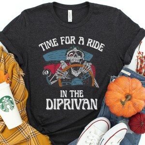 May include: A dark gray t-shirt with a graphic of a skeleton driving a car. The text on the shirt reads "Time for a ride in the dip-rivan".