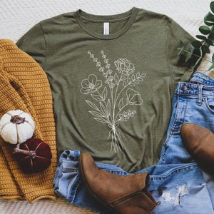 K&ouml;nnte beinhalten: Ein gr&uuml;nes T-Shirt mit einer wei&szlig;en Umrandung eines Blumenstrau&szlig;es. Das T-Shirt liegt auf einer wei&szlig;en, flauschigen Oberfl&auml;che mit einem braunen Pullover, einer blauen Jeans und braunen Stiefeln.