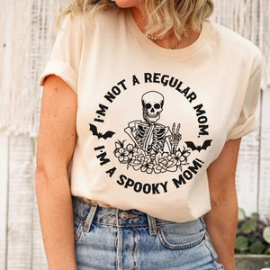 May include: A beige t-shirt with a black graphic of a skeleton holding a peace sign surrounded by flowers and bats. The text on the shirt reads "I'm not a regular mom. I'm a spooky mom!"