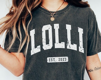 May include: A dark gray t-shirt with the word "LOLLI" printed in white with a distressed look. The shirt also has the text "EST. 2023" printed in white below the word "LOLLI".
