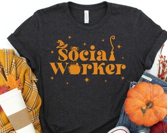 May include: Dark grey heather t-shirt with a graphic of a witch's hat, cauldron, and broom with the text "Social Worker" in orange.