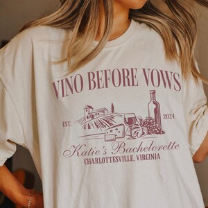 May include: A white t-shirt with a burgundy graphic design that says "Vino Before Vows" with a wine bottle, grapes, cheese, and a glass of wine. The graphic also says "Est. 2024" and "Katie's Bachelorette Charlottesville, Virginia".