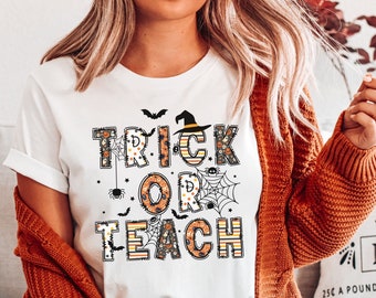 Puede incluir: Camiseta blanca con el texto "TRICK OR TEACH" en naranja, negro y blanco, decorada con murciélagos, arañas y telarañas. La camiseta tiene un sombrero de bruja encima de la palabra "TRICK".