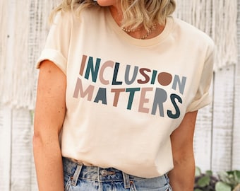 May include: Cream-colored t-shirt with the words "INCLUSION MATTERS" in a colorful, block letter design. The shirt is paired with blue jeans. The background features a white wooden wall and a macrame wall hanging.