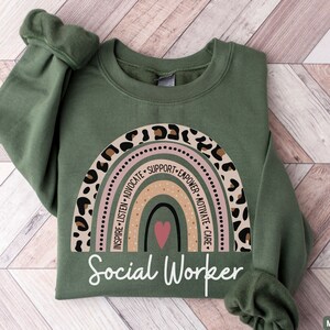 May include: A green sweatshirt with a rainbow design and the words "Social Worker" printed on the front. The rainbow is made up of different colored stripes and has a leopard print border. The words "Inspire, Listen, Advocate, Support, Empower, Motivate, Care" are written around the rainbow.