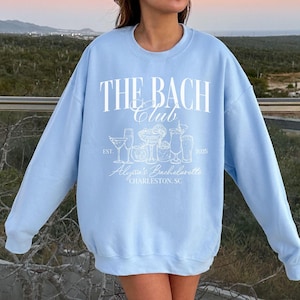 May include: Light blue sweatshirt with white text that reads "The Bach Club Est. 2023 Alyssa's Bachelorette Charleston, SC". The text is surrounded by a graphic of cocktails.