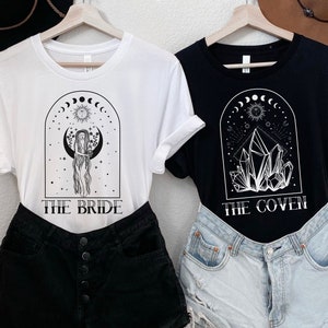 May include: Two t-shirts with a black and white design. The white t-shirt has a woman standing in a crescent moon with the text "The Bride". The black t-shirt has crystals in a crescent moon with the text "The Coven".