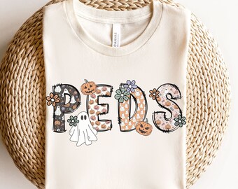 May include: A cream-colored t-shirt with the word "PEDS" spelled out in black outlined letters. Each letter is filled with a pattern of ghosts, pumpkins, and flowers. The shirt is on a woven straw surface.