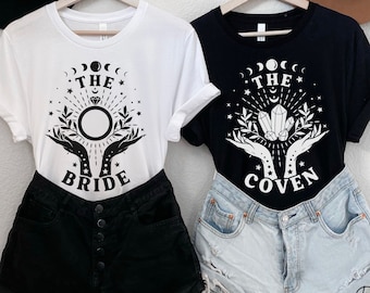 以下が含まれることがあります： 黒い文字とグラフィックが描かれた白いTシャツ2枚。左側のシャツには「The Bride」とリングと手が描かれている。右側のシャツには「The Coven」とクリスタルと手が描かれている。