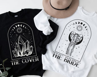 May include: Two black and white sweatshirts with a celestial design. One sweatshirt says "The Coven" and features a crystal cluster. The other sweatshirt says "The Bride" and features a woman with long hair standing in a crescent moon.