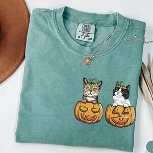 May include: A light green t-shirt featuring a graphic of two cats wearing pumpkin hats and sitting inside pumpkins. The tag reads "COMFORT COLORS".