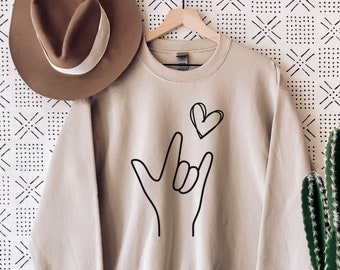 May include: A beige sweatshirt with a black hand sign design and a small heart above it. The hand sign is the American Sign Language symbol for "I love you".
