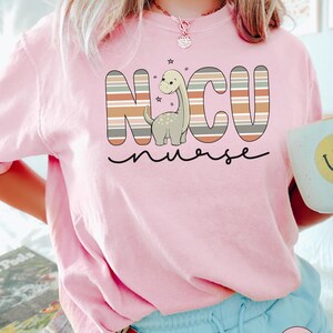 May include: Pink t-shirt with a striped graphic that says "NCU nurse" with a cartoon dinosaur in the middle.