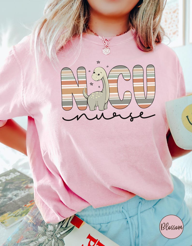 May include: Pink t-shirt with a striped graphic that says "NCU nurse" with a cartoon dinosaur in the middle.