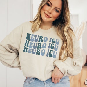 Pode incluir: Uma camisa de moletom de cor creme com as palavras "Neuro ICU" repetidas tr&ecirc;s vezes em uma fonte retr&ocirc; azul e verde.