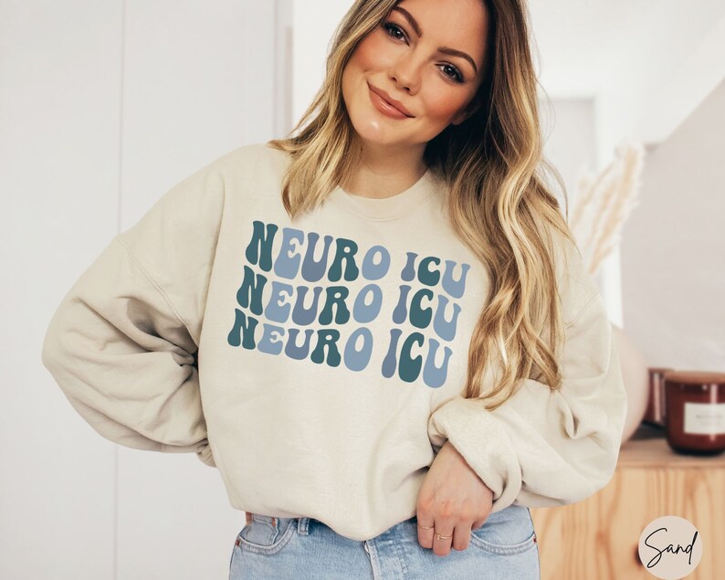 Pode incluir: Uma camisa de moletom de cor creme com as palavras "Neuro ICU" repetidas tr&ecirc;s vezes em uma fonte retr&ocirc; azul e verde.