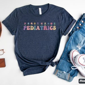 Puede incluir: Una camiseta azul marino jaspeada con un gr&aacute;fico de flores de colores y la palabra "PEDIATRICS" en letras arco&iacute;ris.