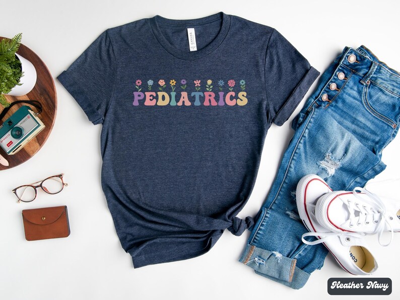 Puede incluir: Una camiseta azul marino jaspeada con un gr&aacute;fico de flores de colores y la palabra "PEDIATRICS" en letras arco&iacute;ris.