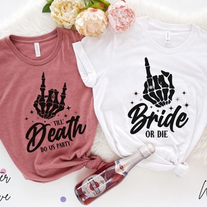 Puede incluir: Dos camisetas con una mano de esqueleto haciendo el dedo medio. La camiseta de la izquierda es de color malva jaspeado y dice "Till Death Do Us Party". La camiseta de la derecha es blanca y dice "Bride or Die".