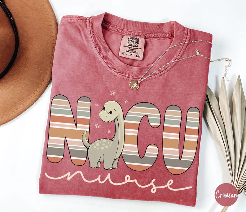 May include: A red t-shirt with a striped design and a cartoon dinosaur. The shirt says "NCU nurse" in a pink script font.