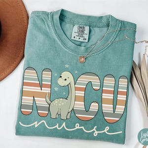 May include: A teal blue t-shirt with a striped design that spells out "NCU nurse" with a cartoon dinosaur in the middle.