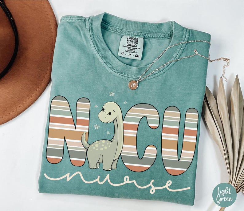 May include: A teal blue t-shirt with a striped design that spells out "NCU nurse" with a cartoon dinosaur in the middle.