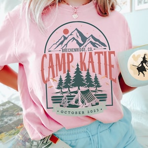 May include: Pink t-shirt with a graphic design of a mountain range, a tent, and trees. The text "Breckenridge, CO." and "Camp Katie" are printed on the shirt. The text "October 2023" is printed below the graphic.