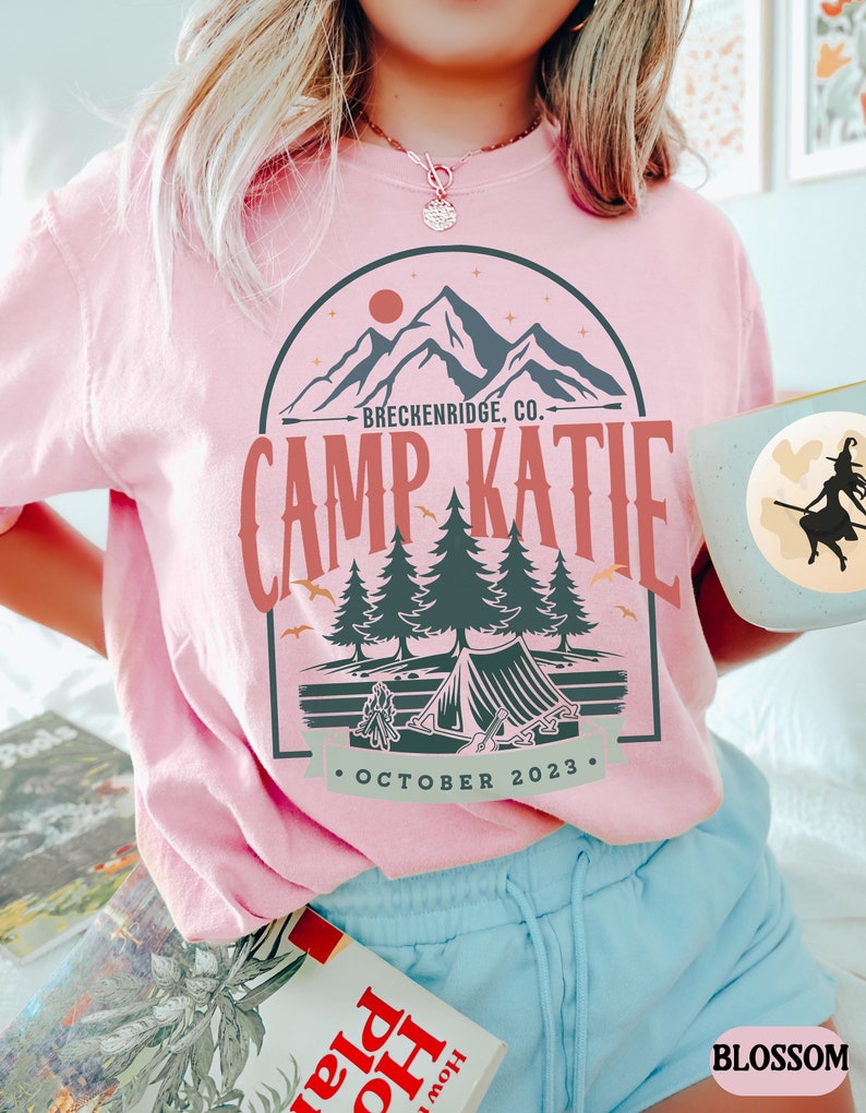 May include: Pink t-shirt with a graphic design of a mountain range, a tent, and trees. The text "Breckenridge, CO." and "Camp Katie" are printed on the shirt. The text "October 2023" is printed below the graphic.