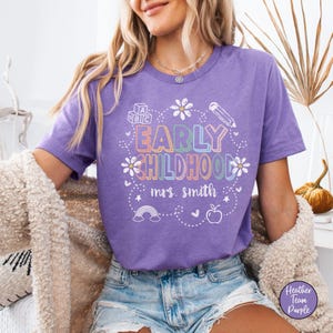 Pode incluir: Uma camiseta roxa com as palavras "EARLY CHILDHOOD" em letras coloridas, juntamente com o nome "mrs. smith". A camiseta apresenta um l&aacute;pis, flores, um arco-&iacute;ris e um design de ma&ccedil;&atilde;. A camiseta &eacute; feita de um material macio.